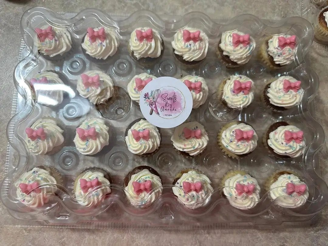 Two dozen mini cupcakes with swirled vanilla buttercream, pink fondant bows and sprinkles, boxed with a Sweets By Amanda sticker