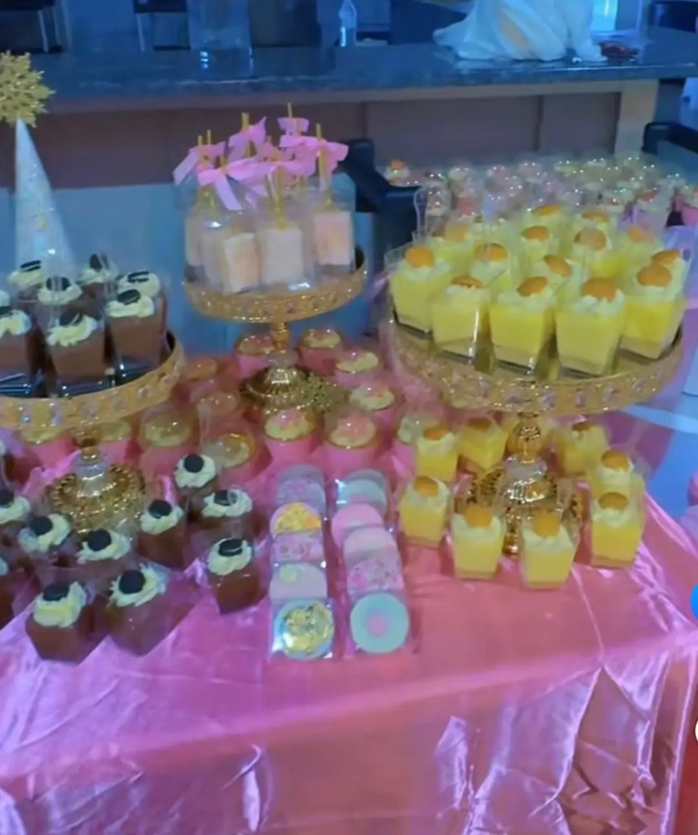 Custom dessert table on pink linen with gold tiered stands holding mousse cups, cupcakes, chocolate-dipped treats and decorated cookies