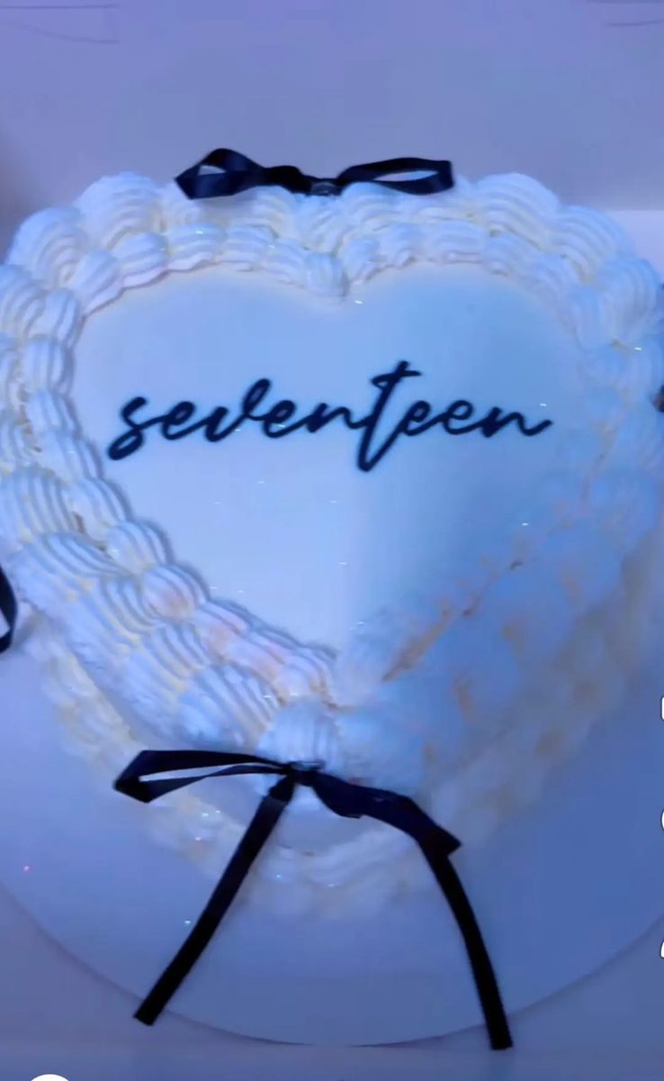 Heart-shaped cake in white buttercream with a piped shell border, black satin bows and a script topper reading seventeen