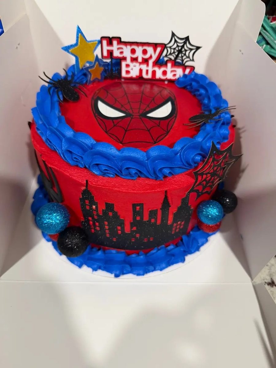 Tall red buttercream birthday cake with a Spider-Man mask on top, a piped blue rosette border, a black city-skyline cut-out around the side and a Happy Birthday topper
